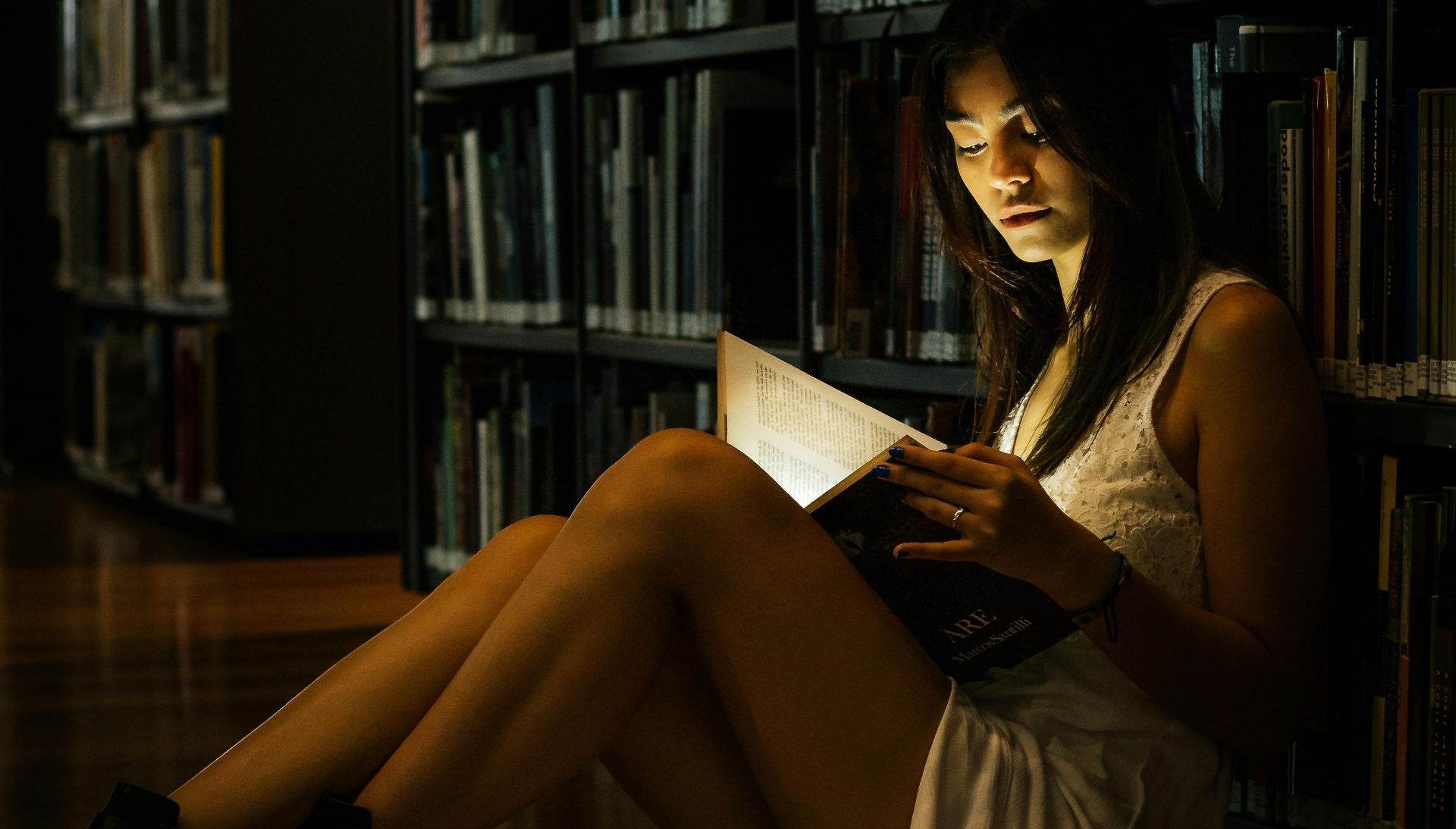 A woman reads a glowing book on the floor of a dimly lit library, creating a magical atmosphere.