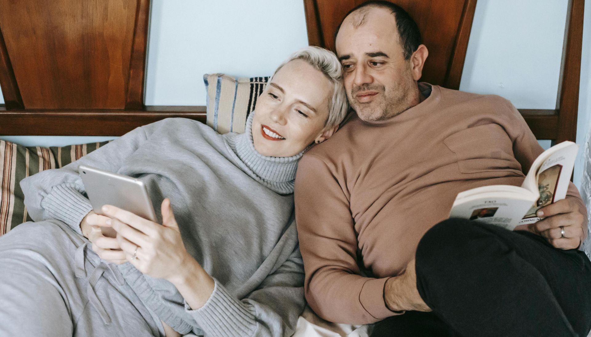 High angle of happy adult woman using tablet and cuddling with ethnic husband reading interesting book on bed at home
